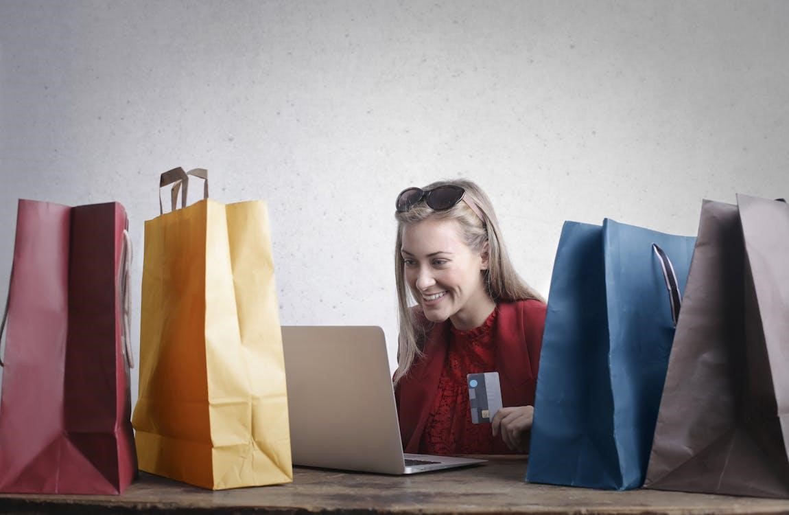 A Woman Holds a Credit Card While Smiling at Her Laptop Screen While Surrounded by Shopping Bags